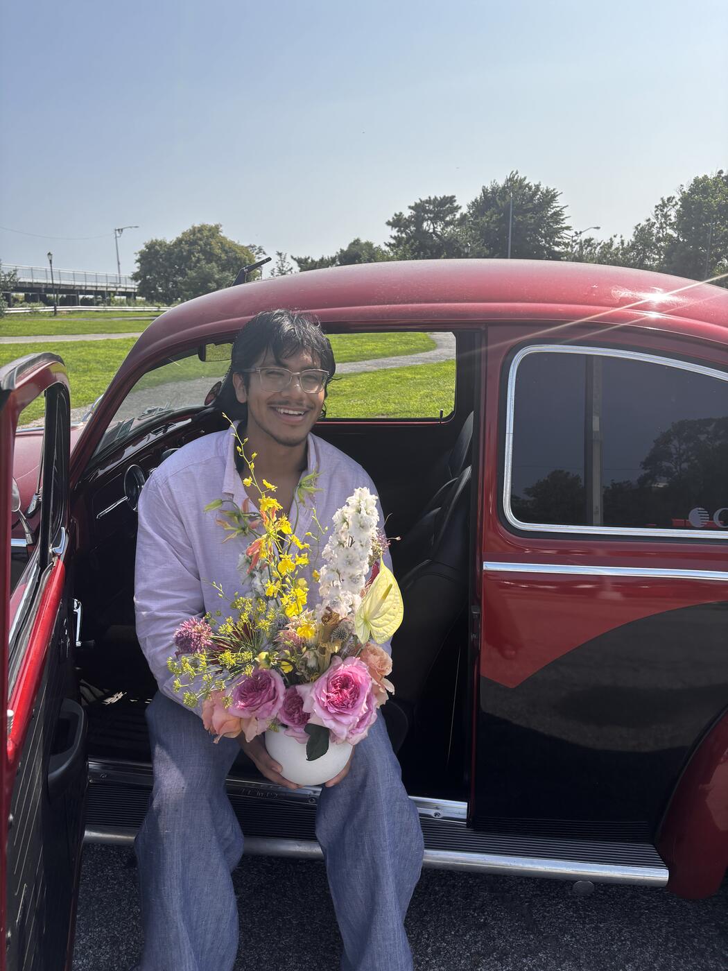 Hern seated in the doorway of a vintage car, holding an arrangement of garden roses, oncidium orchids, and anthurium